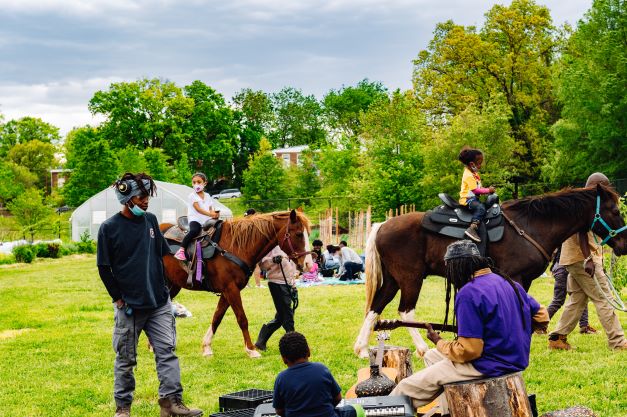 Photo of Dreaming Out Loud's 2021 Spring Fest depicting people outside on an urban farm, with a few riding horses and others playing music