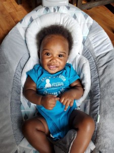 Top-down photo of a smiling baby lying on a white bed who is wearing a blue onesie