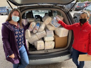 Photo of two people wearing masks and posing for a camera by the open trunk of a car, which is full with packs of diapers