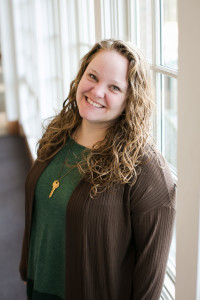 Photo of a person with long, curly blonde hair smiling at the camera wearing a dark green jacket, green top, and a necklace with a key on it