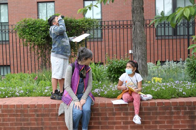 Photo of two young students outside with an adult. One is using binoculars to look up while another is seated next to the adult and having a conversation