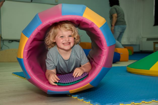 Photo of a young child smiling for the camera while posing inside a multi-colored barrel