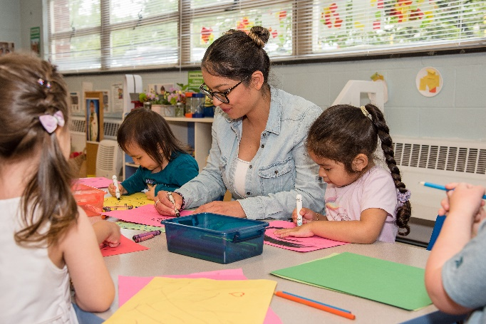 Photo of a teacher wearing glasses and a denim jacket coloring with young students in a classroom