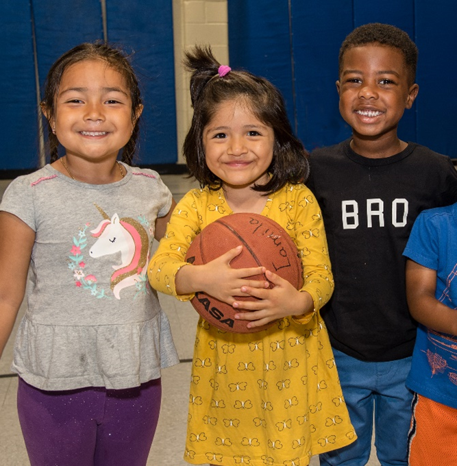 Photo of three young children smiling and posing for the camera. The child on the left is wearing a gray shirt with a colorful unicorn on it. The child in the middle is wearing a yellow printed dress, holding a basketball. And the child on the right is wearing a black t-shirt with the word "BRO" on it.