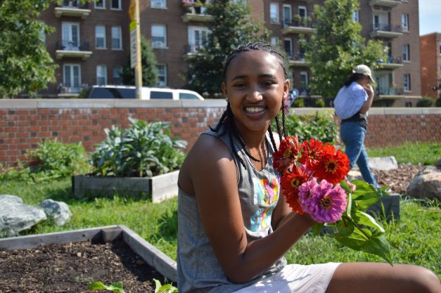 A person wearing braids and a gray tank top holding a bunch of red and pink flowers, smiling at the camera in a garden