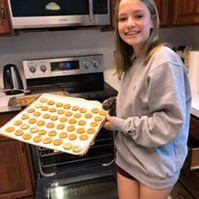 A young volunteer with shoulder-length blonde hair wearing a gray sweater smiling at the camera with homemade dog treats baked for Homeward Trails Adoption Center