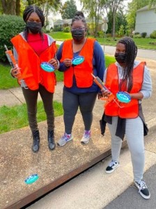 Three volunteers, all wearing bright orange vests and black face masks, posing for the camera on the sidewalk with clipboards in hand