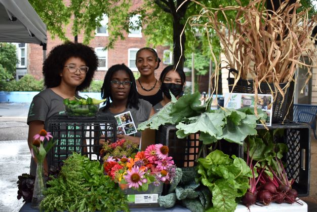 A group of high school Mighty Greens youth staff posing for the camera by their booth at the farmers market, which is filled with fresh produce
