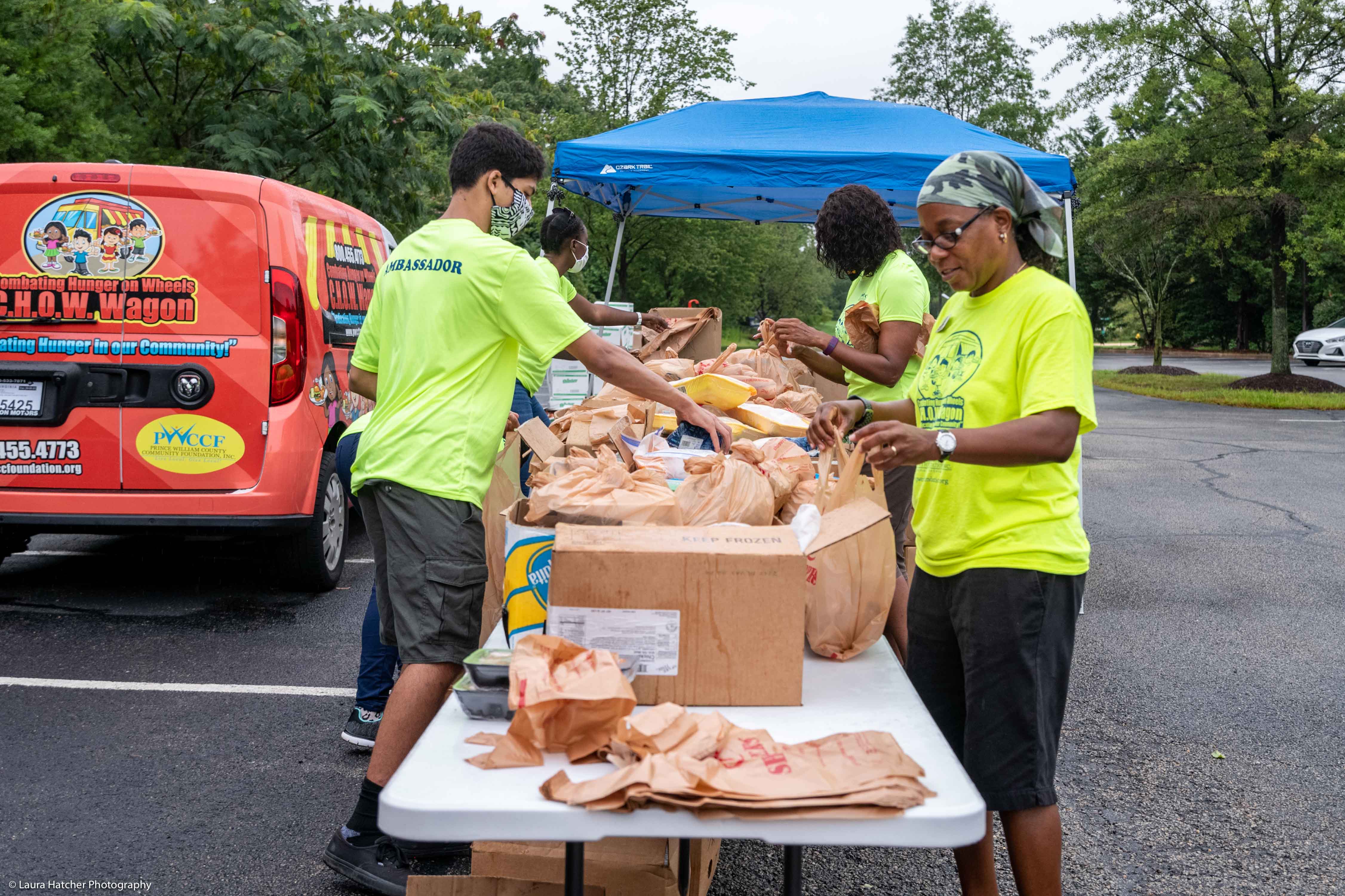 A group of volunteers wearing neon green shirts sorting boxes of food on a table set up in a parking lot, next to a blue makeshift tent and the C.H.O.W. Wagon