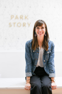 Photo of Meghan Evans, owner of Park Story, a woman with long brown hair wearing lipstick and dangly earrings, smiling at the camera, in a denim jacket, white top, and black pants.