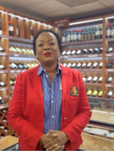 Photo of Eveline Ngassa, owner of Wine & Organic, a woman with short black hair wearing red lipstick and a red blazer, posing for the camera, in a blue striped button-down.