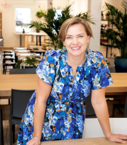 Photo of Melina Selimbegovic, owner of Artsy Beast, a woman with brown-blonde hair wearing dangly earrings, smiling at the camera, in a patterned blue floral dress.