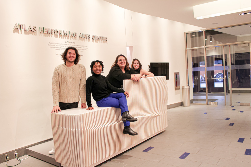 Photo taken in lobby of the Atlas Performing Arts Center with an accessible welcome desk created by artist Salvator Pirrone, on top of which four Atlas community members are seated, smiling for the camera.