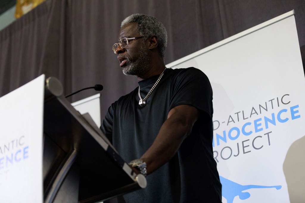 MAIP client Ransom Watkins shares his story, speaking at a podium. He is a Black man with short black and white hair, wearing glasses, a black tee, and a necklace.