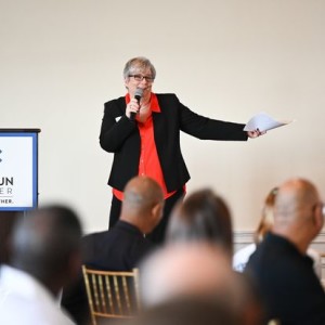 Loudoun Cares Executive Director Valerie Pisierra, wearing a pair of glasses, black blazer, and red top, speaking in front of a crowd