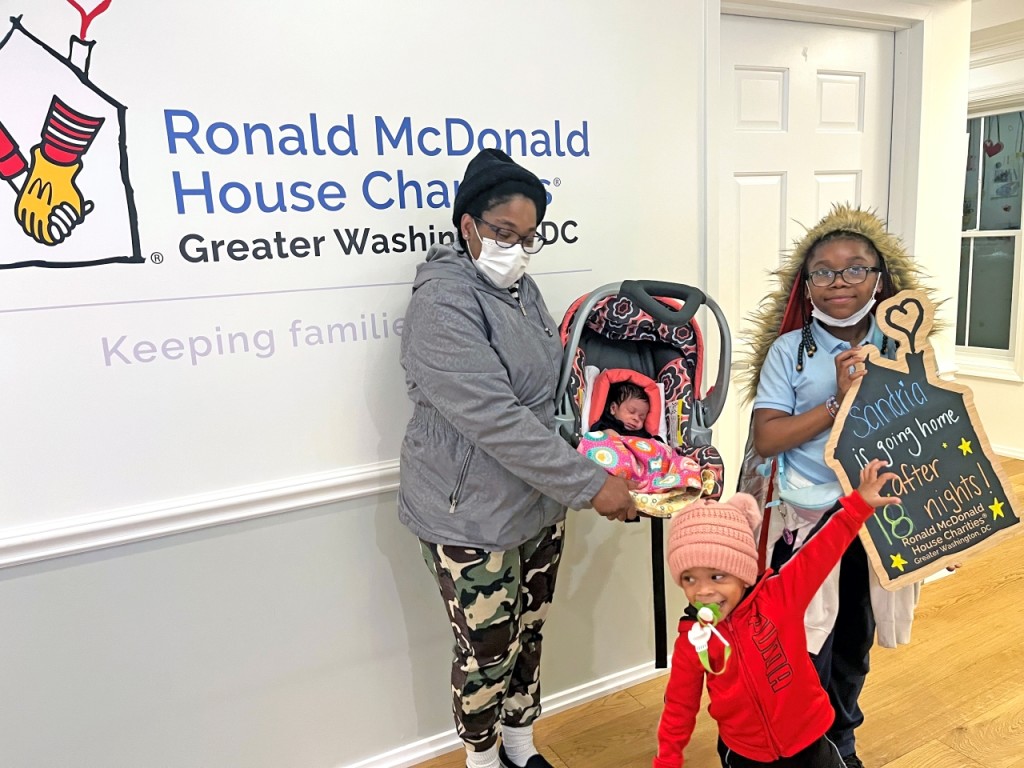 Photo of a mother posing with her three young children by the Ronald McDonald House Charities Greater Washington, DC sign, all of whom are smiling at the camera