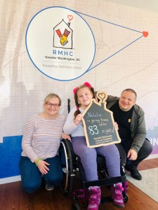 Parents posing next to their daughter by the Ronald McDonald House Charities of Greater Washington, DC sign. The daughter, Natalia, is sitting in a wheelchair and holding a chalkboard sign that reads "Natalia is going home after 83 nights."