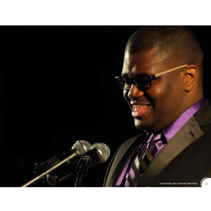 Photo of Christian Souvenir, a young Black man who is neurodivergent and participated in BroadFutures' internship program. He is wearing glasses, a suit and tie with a purple shirt, and is speaking into a microphone at a podium.